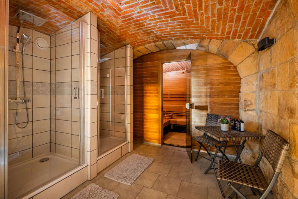 Wide view of the wellness area at Resort Mezná featuring two glass-enclosed showers, the entrance to a wooden sauna and a seating area beneath a brick vaulted ceiling.